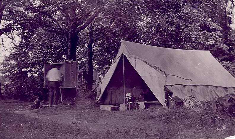 Black & white photograph of Camp Shady Shore.  Picture of unidentified man in a 3/4 enclosure that is attached to a tree. Partial view inside open tent.
