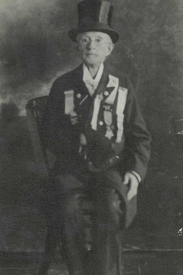 Black & white photograph of an older Dr. Walker.  She is seated on a wooden chair.  Dressed in trousers, jacket, white shirt and top hat.  Her jacket is decorated with ribbons, pins and the Congressional Medal of Honor.   Dr. Walker is wearing eyeglasses and is gazing at the camera.
